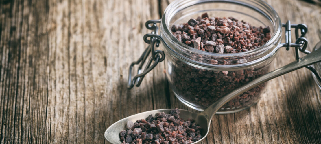 Ein Glass und ein Metalllöffel gefüllt mit schwarzem Salz. Auf Holztisch