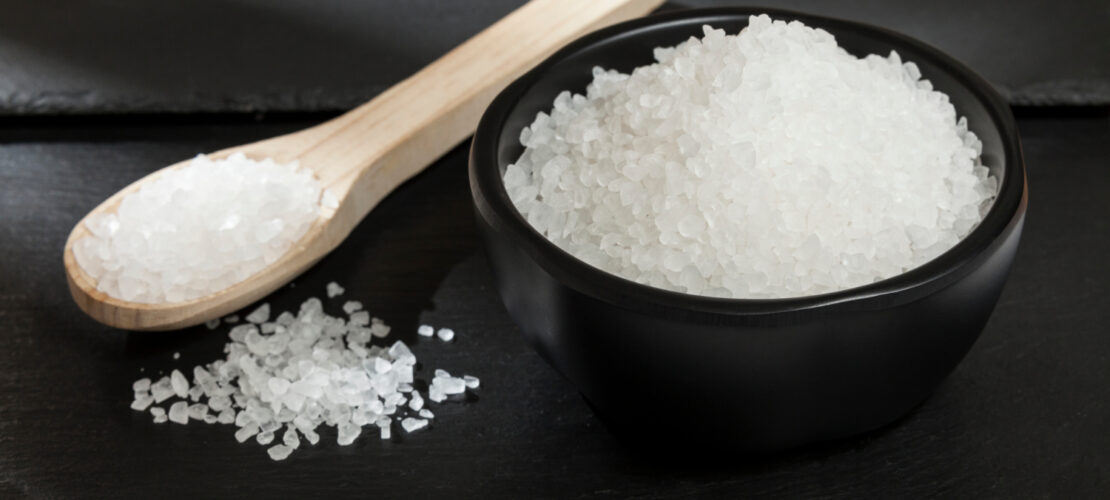 Eine Schwarze Schüssel und ein Heller Holzlöffel Vor dunklen Hintergrund. Die Schüssel und der Löffel sind mit Weißem Salz gefüllt