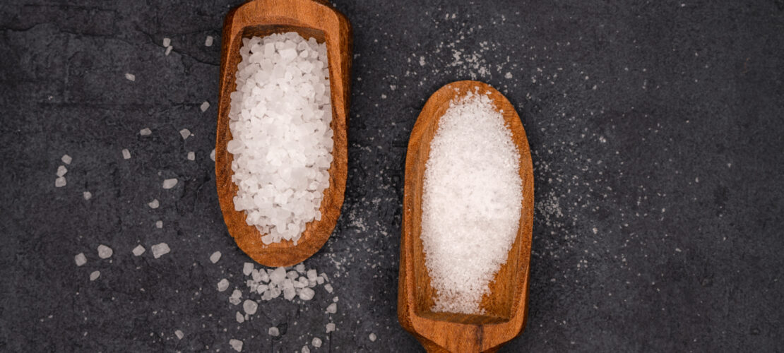 zwei Hollöffel liegen gegenüber beide sind mit Salz gefüllt. Einer mit feinem salz der Andere mit Groben Salz. Auf dunklem Hintergrund